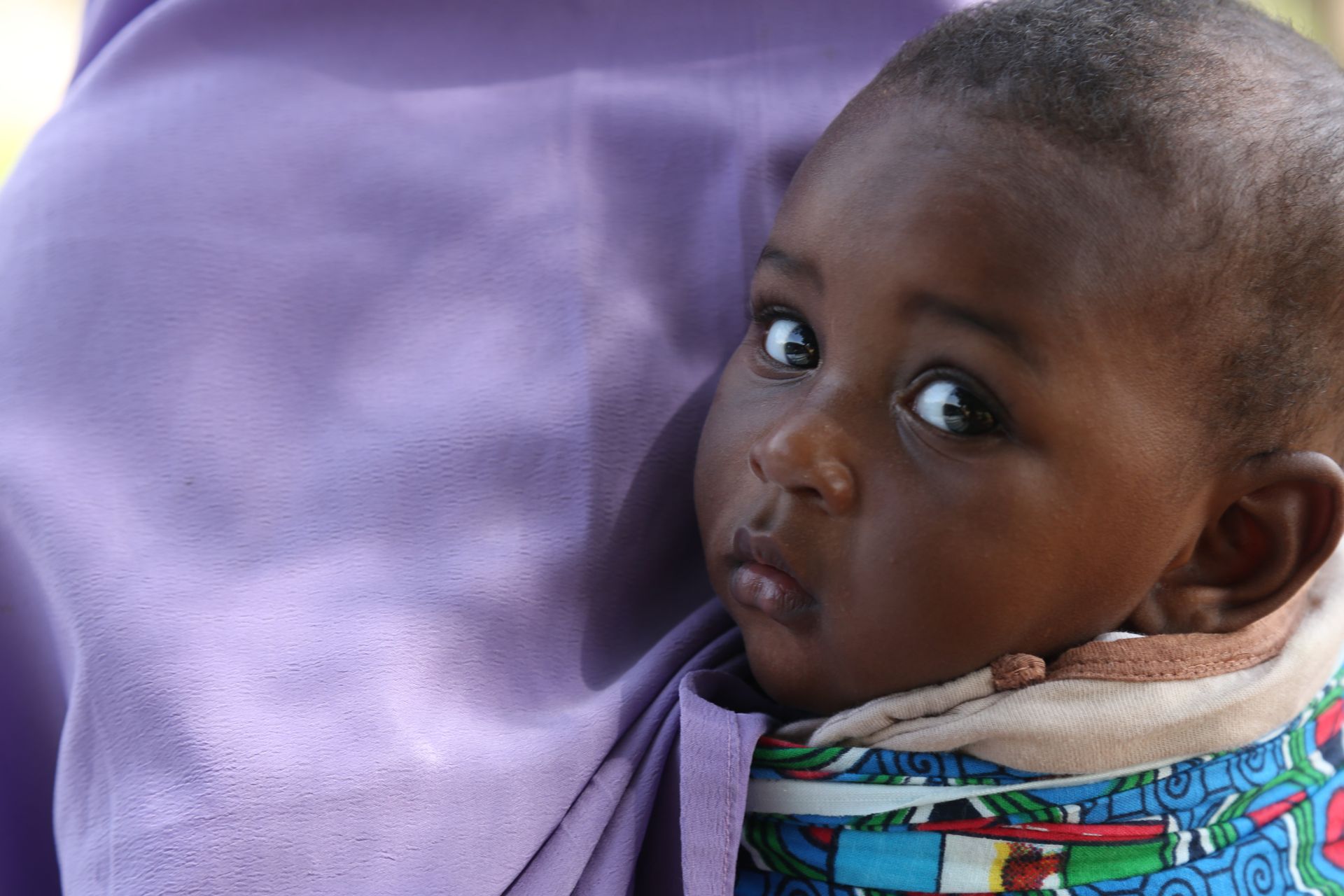 A young child with dark skin peers out from a purple cloth, looking directly at the camera.