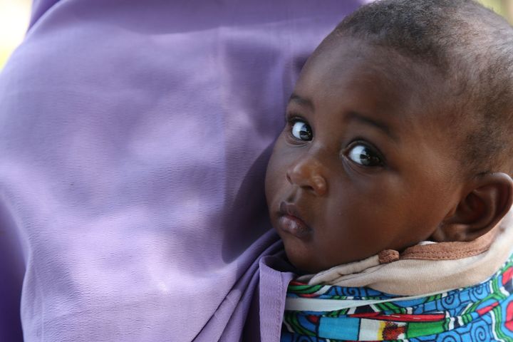 A baby, with dark skin and a serious expression, is held in a purple wrap.