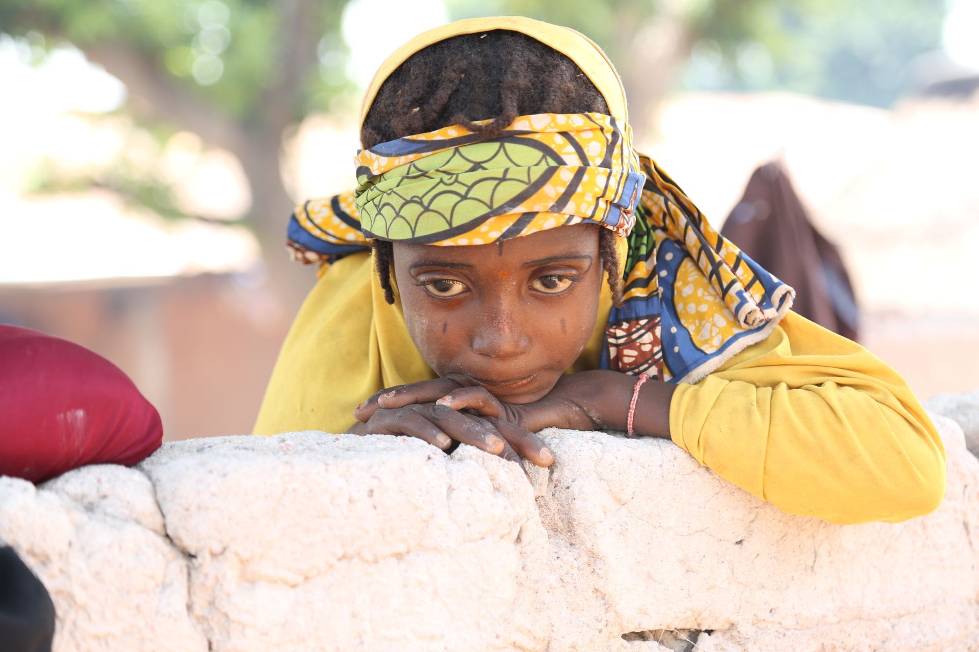 Young girl in yellow headscarf and shirt leans on a brick wall, looking down.