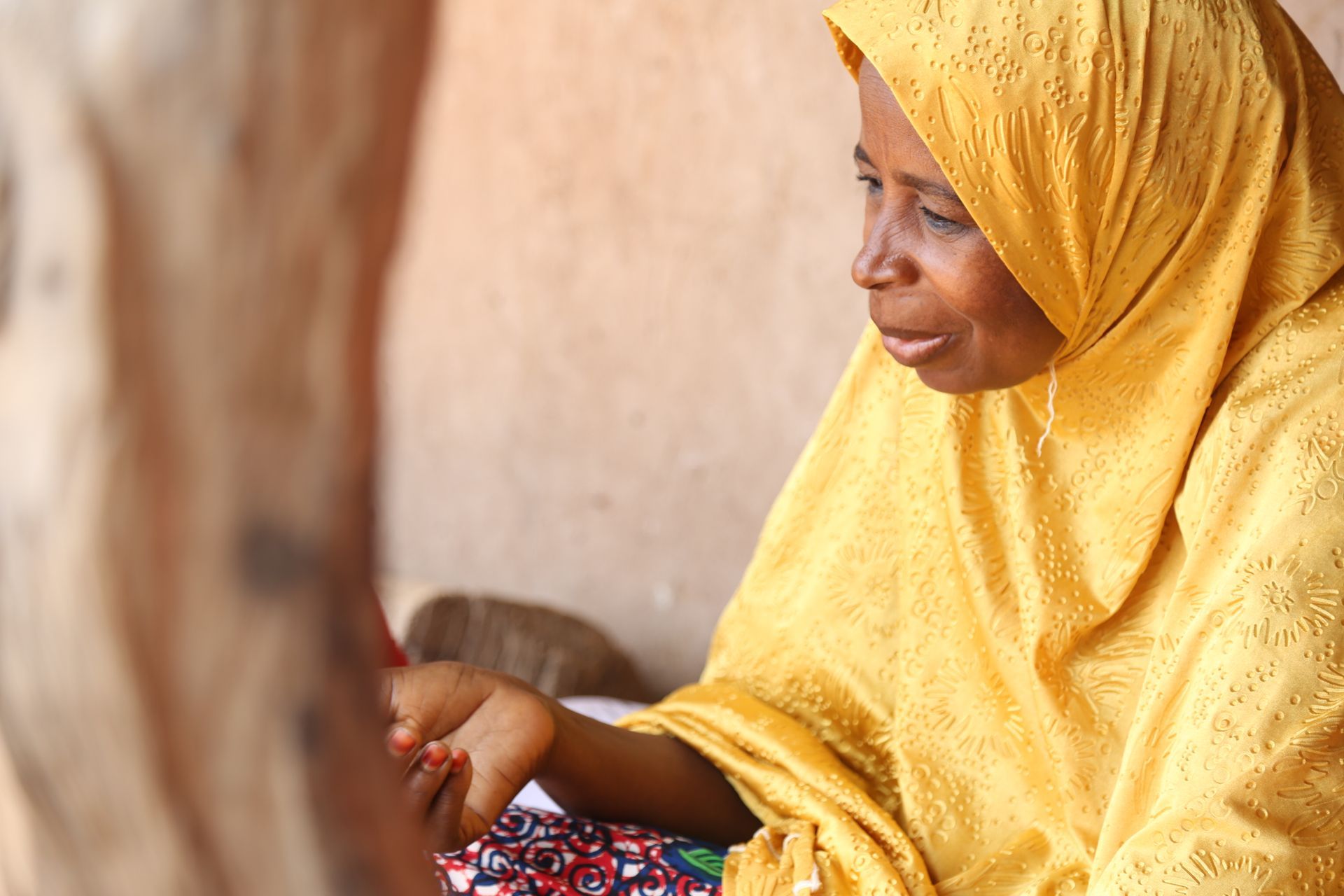 Woman in yellow headscarf seated, hand raised, looking concerned.