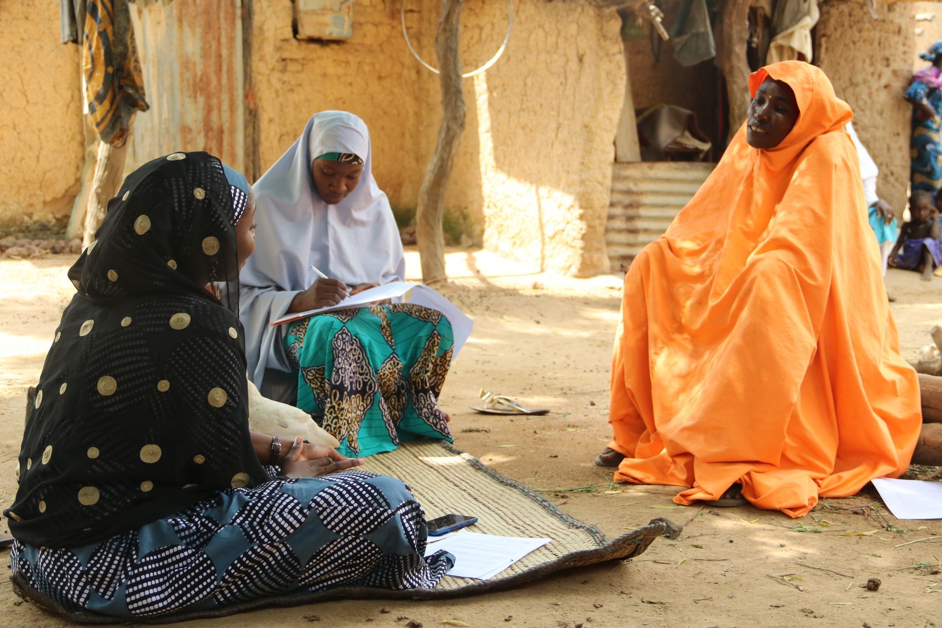 Three women in headscarves sit on mat, one writing, another talking, outdoors in a village.