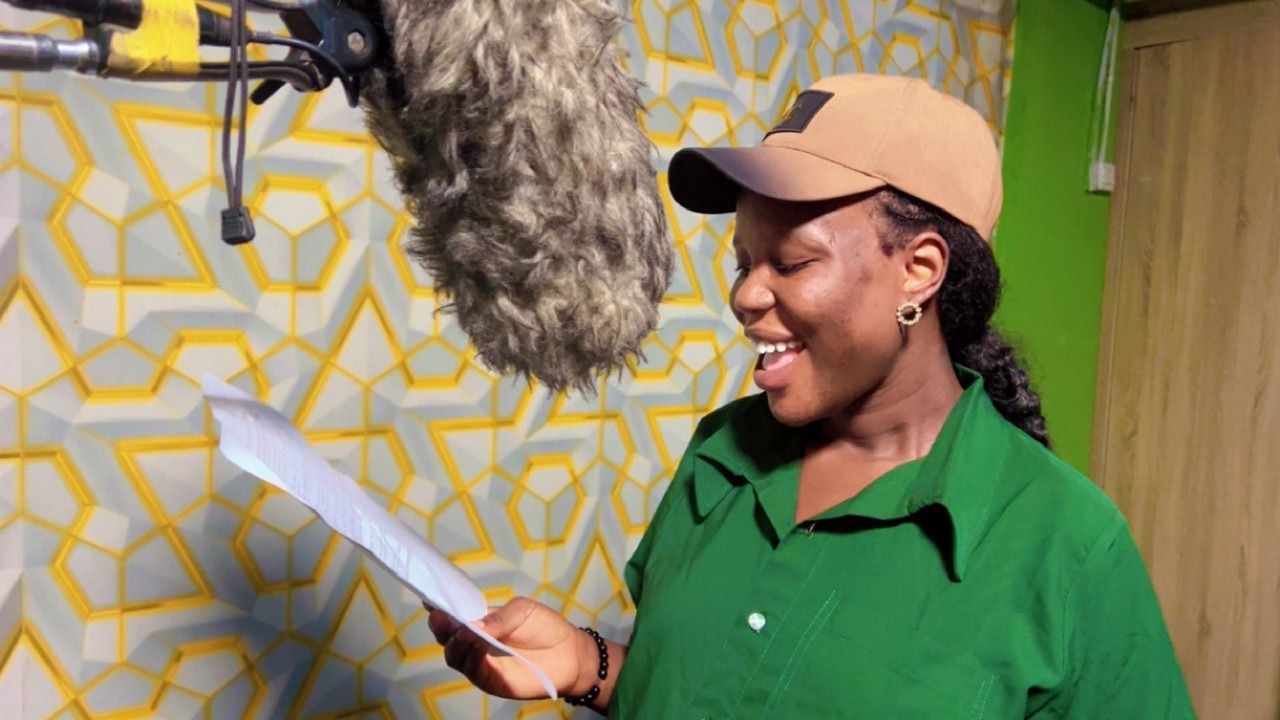 Woman in cap, green shirt, smiling, reading from paper in recording studio with microphone.