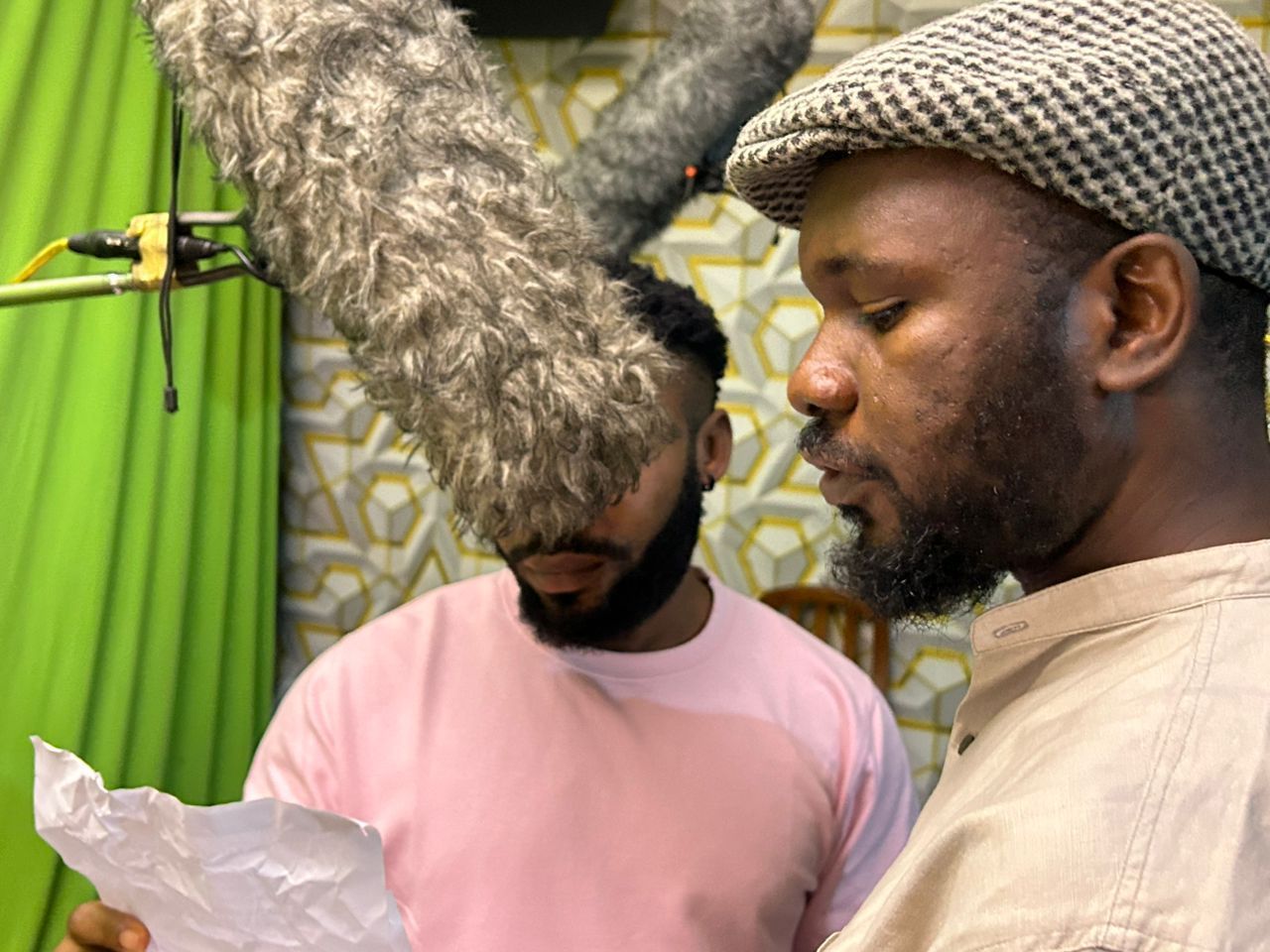 Two men in a studio, reading a script under a microphone. One wears a hat, the other a pink shirt.