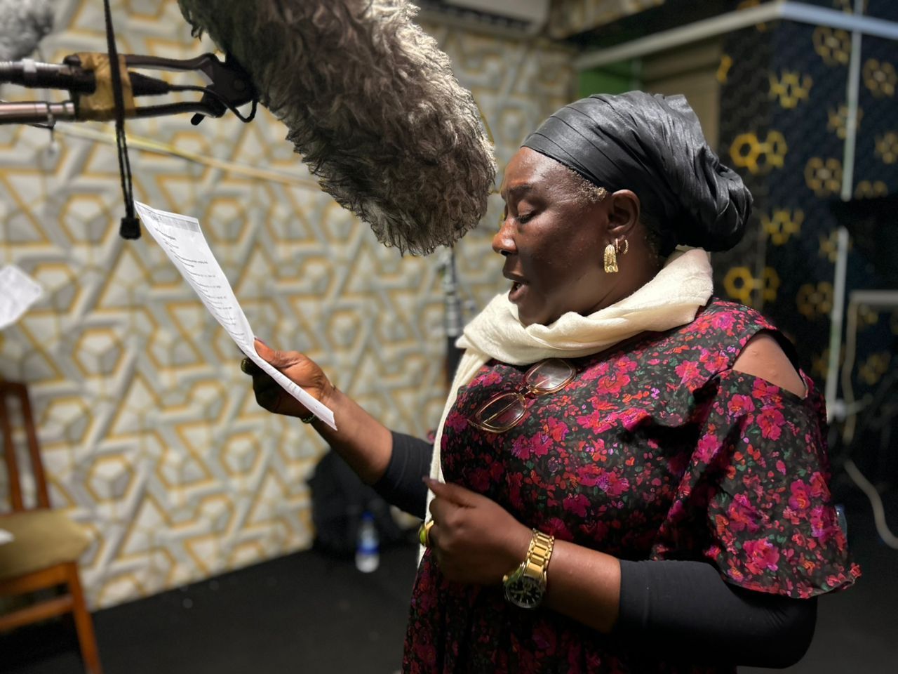 Woman in a recording studio reading a script, wearing a headscarf, and holding a microphone overhead.