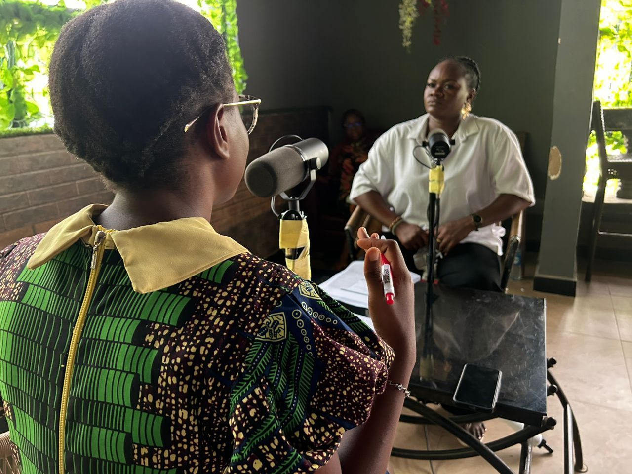 Two women in a podcast setup; one speaking into a mic, the other listening. Outdoors setting.