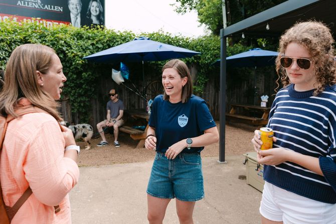 Three people chatting outdoors near blue umbrellas, one holding a drink.