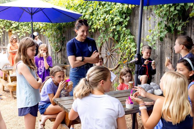 Children gathered around a picnic table outdoors while an adult stands nearby under blue umbrellas