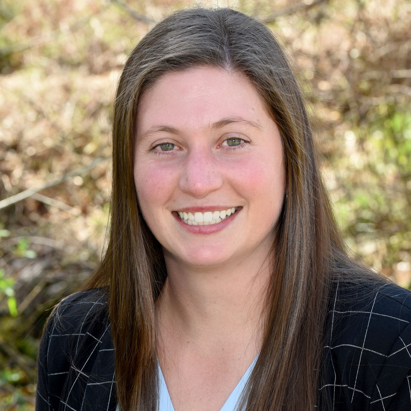 Smiling woman with long brown hair in a black blazer outdoors with blurred foliage background