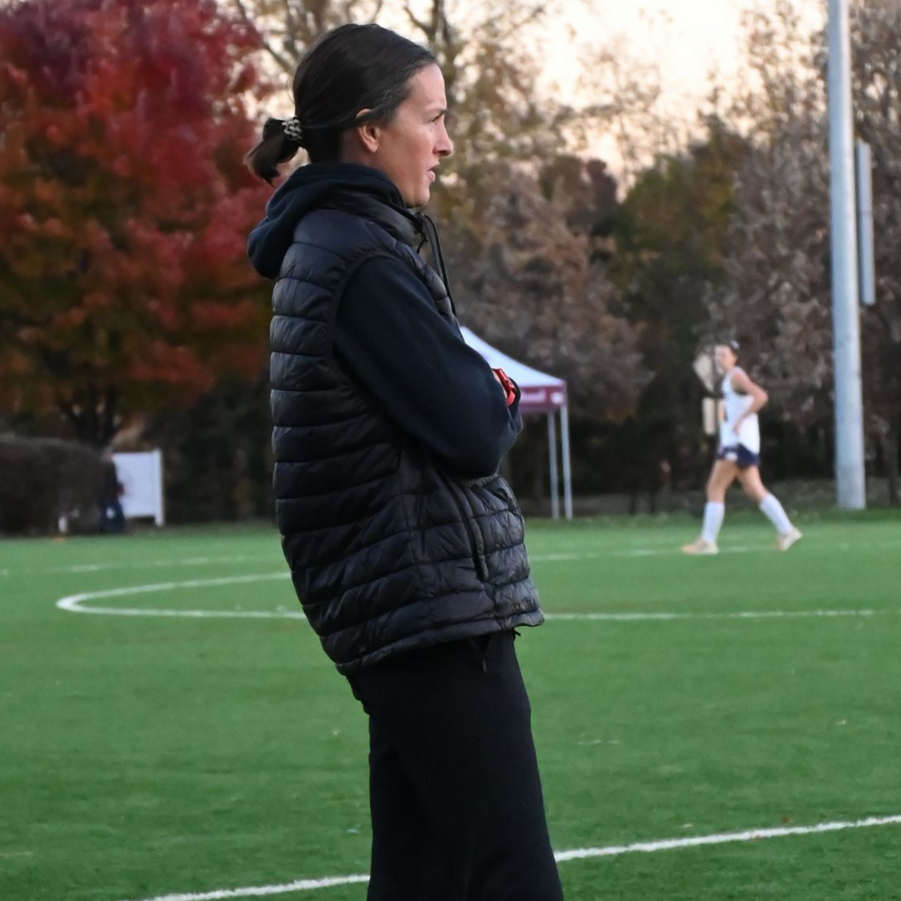 Coach in black jacket watches a soccer game from the sideline on a green field