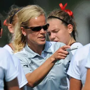 Coach in sunglasses points while speaking to athletes in light blue uniforms outdoors