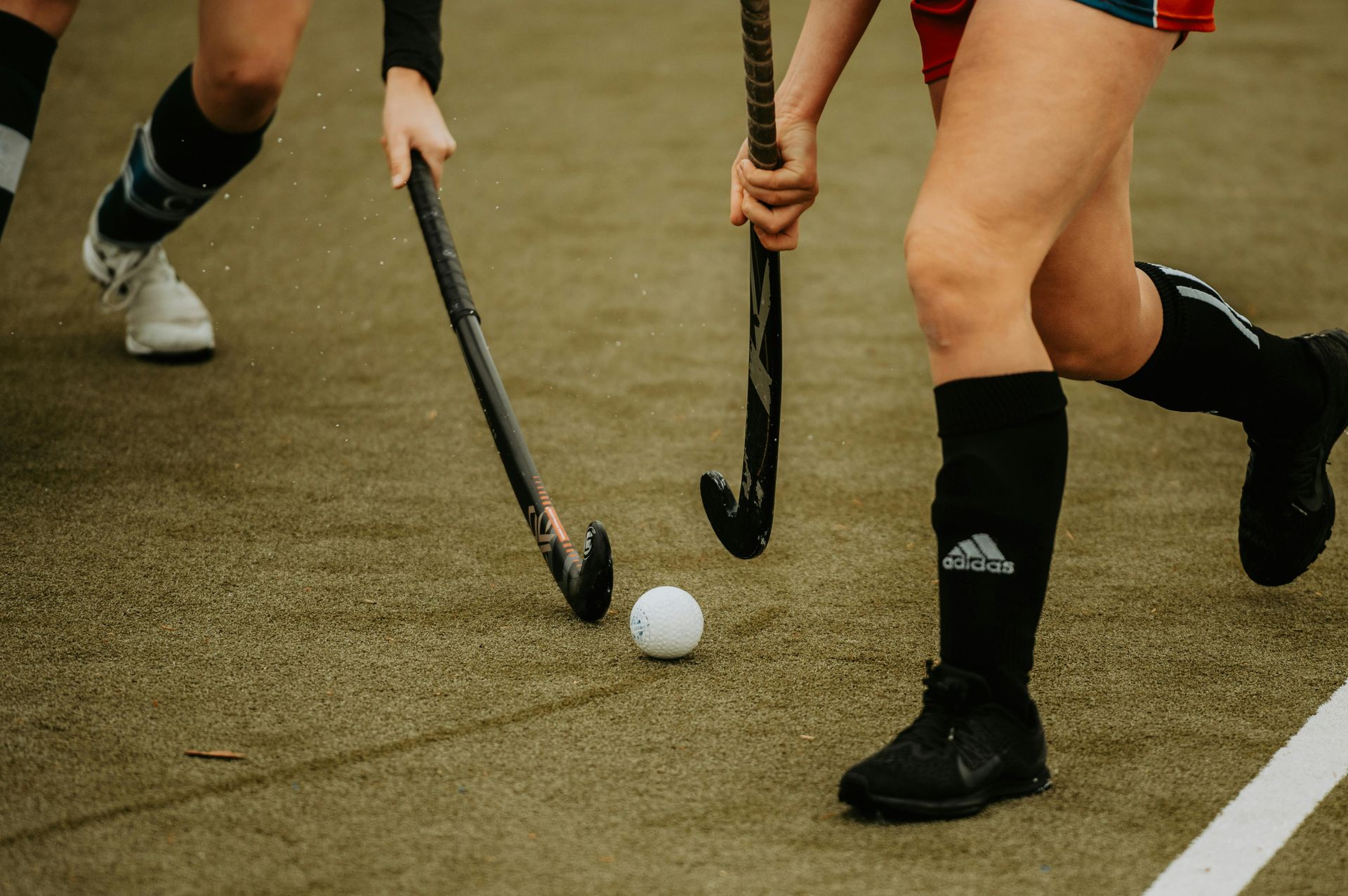 Field hockey players in black socks chase a white ball on a grass pitch.