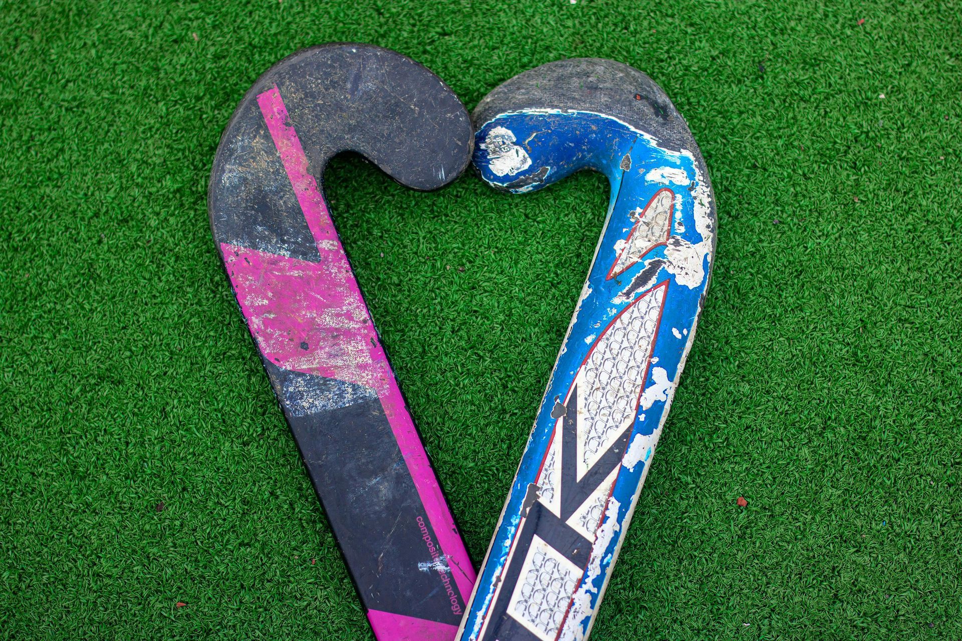 Two overlapping field hockey sticks on green turf, one pink and black, one blue and white.