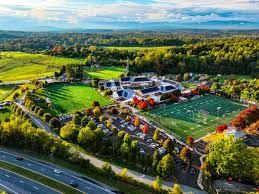 Aerial view of a sports complex with green fields, a running track, parking lot, and surrounding trees.