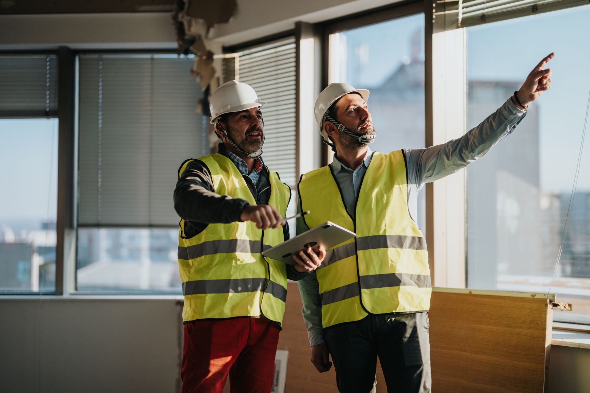 Zwei Bauarbeiter in Sicherheitswesten und Schutzhelmen inspizieren ein Gebäude, einer zeigt nach oben.