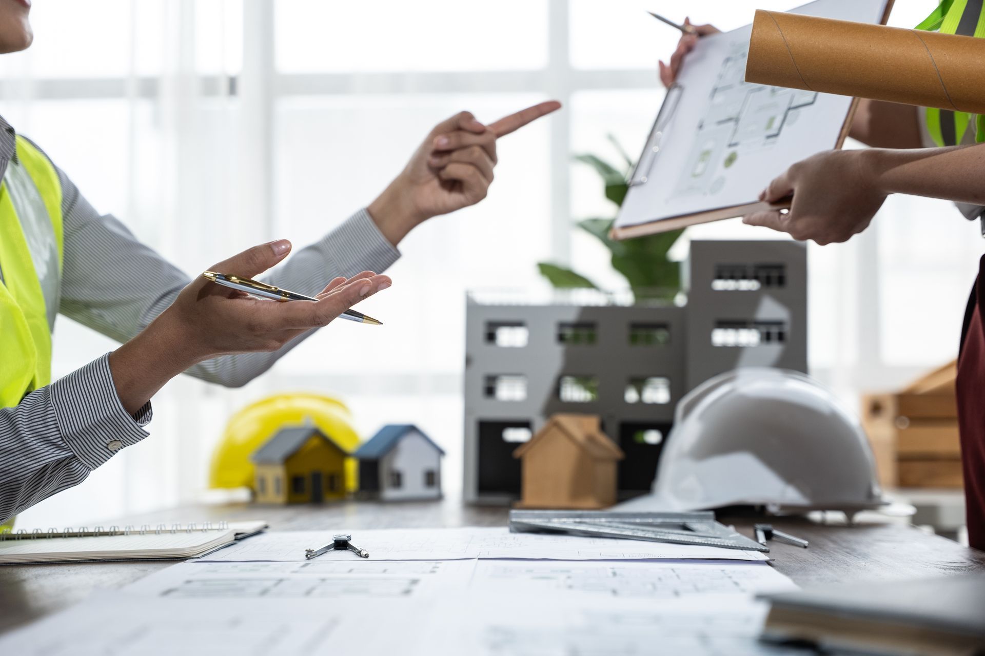 Architekten in Sicherheitswesten besprechen an einem Schreibtisch mit Musterhäusern und Bauplänen Bauentwürfe.