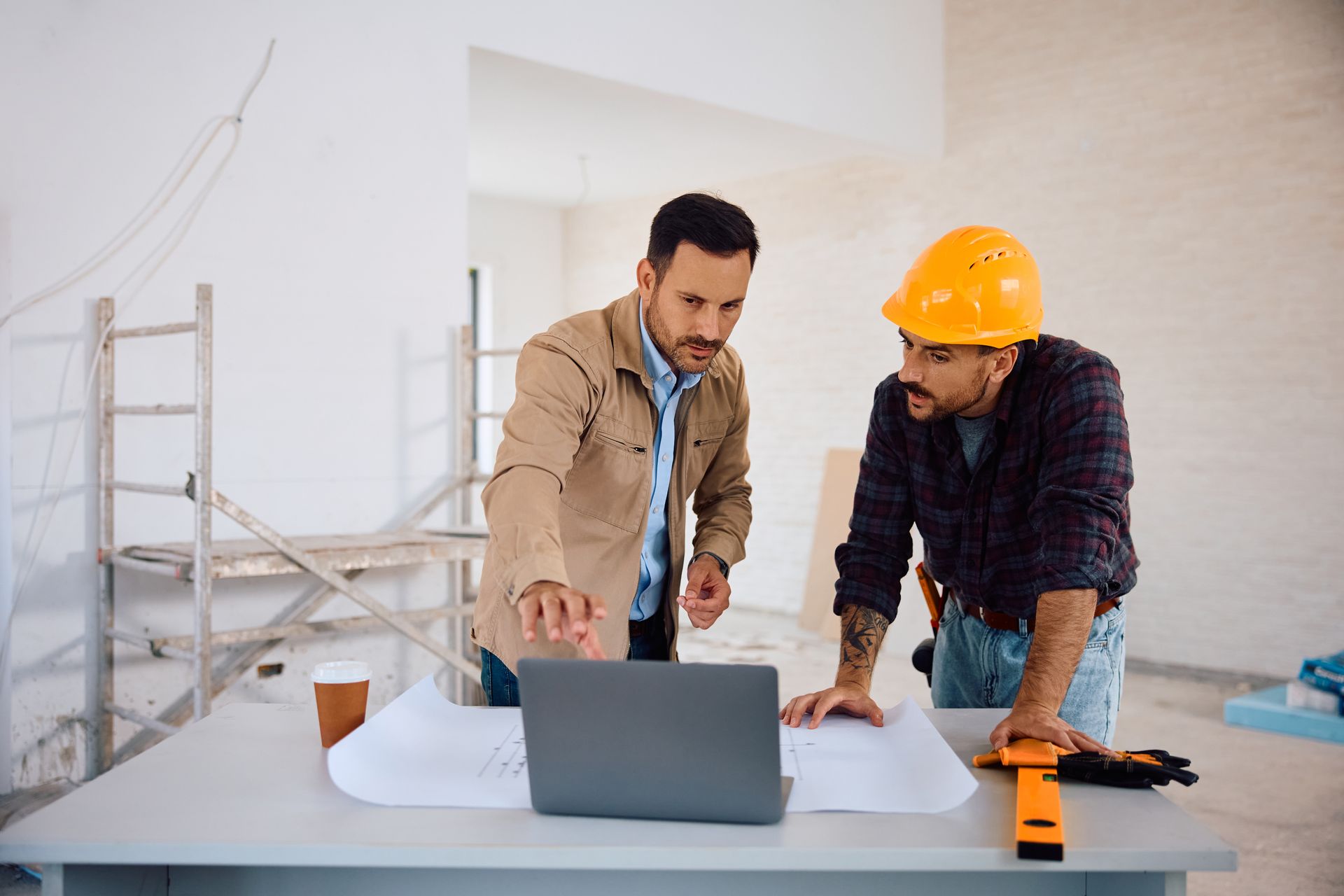Zwei Männer studieren Baupläne und einen Laptop auf einer Baustelle.