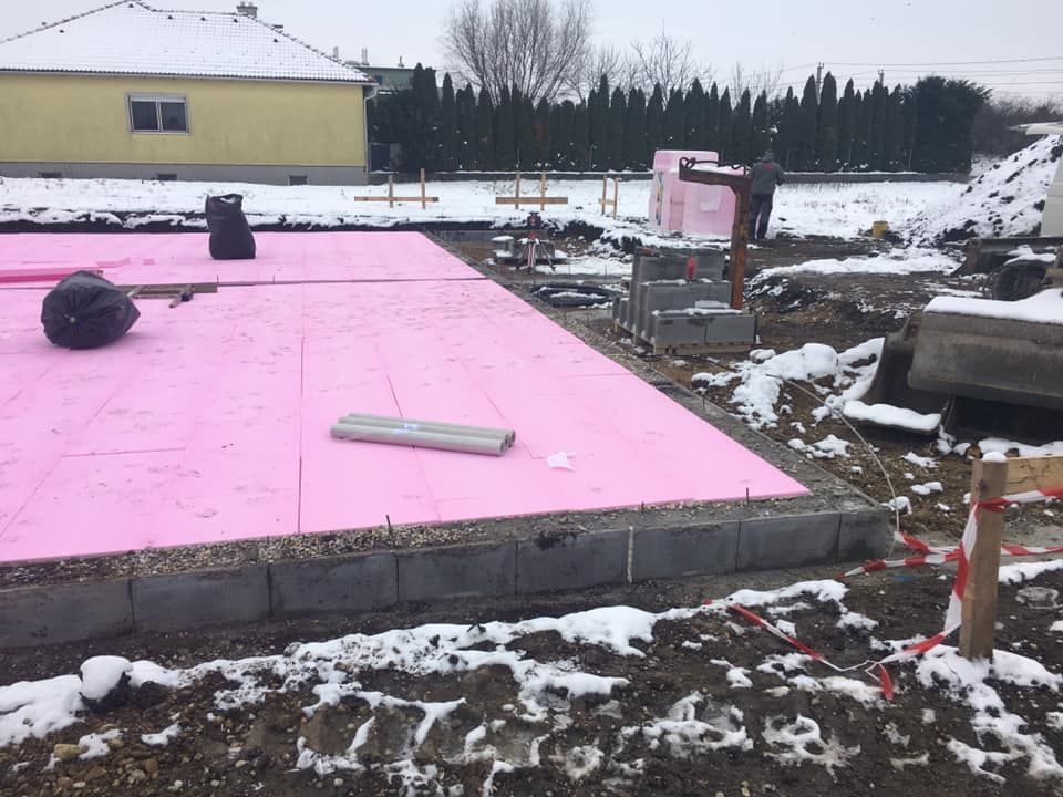 Eine Baustelle mit rosa Dämmung, die ein Fundament bedeckt, Schnee auf dem Boden und eine im Hintergrund arbeitende Person.