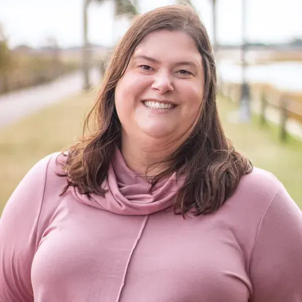 Woman with brown hair smiles, wearing a pink top, outdoors near water and greenery.