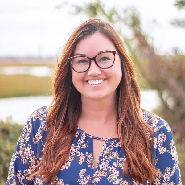 Woman with glasses smiles outdoors, wearing a blue floral top, with long auburn hair.