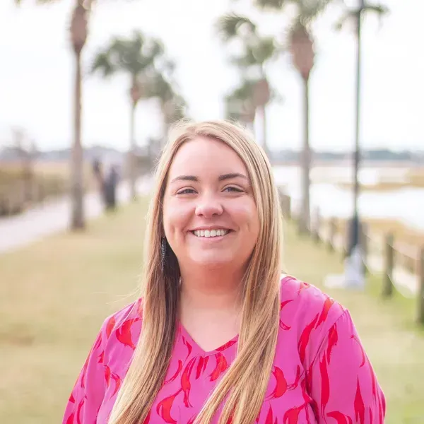 Blond woman in pink top smiles outdoors, palm trees in background.