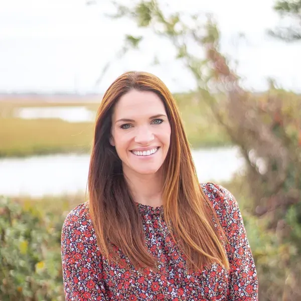 Woman with long auburn hair smiles outdoors, wearing a floral patterned top; blurred background of water and greenery.