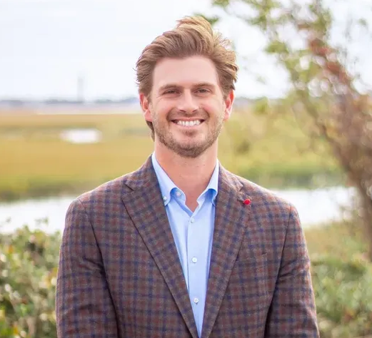 Man in brown plaid blazer smiles outside, blue shirt.