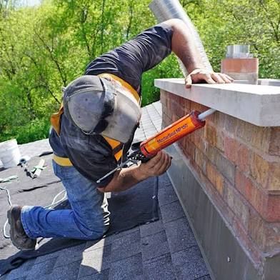 A person kneels on a residential roof, using a caulk gun to seal the edge of a brick chimney.