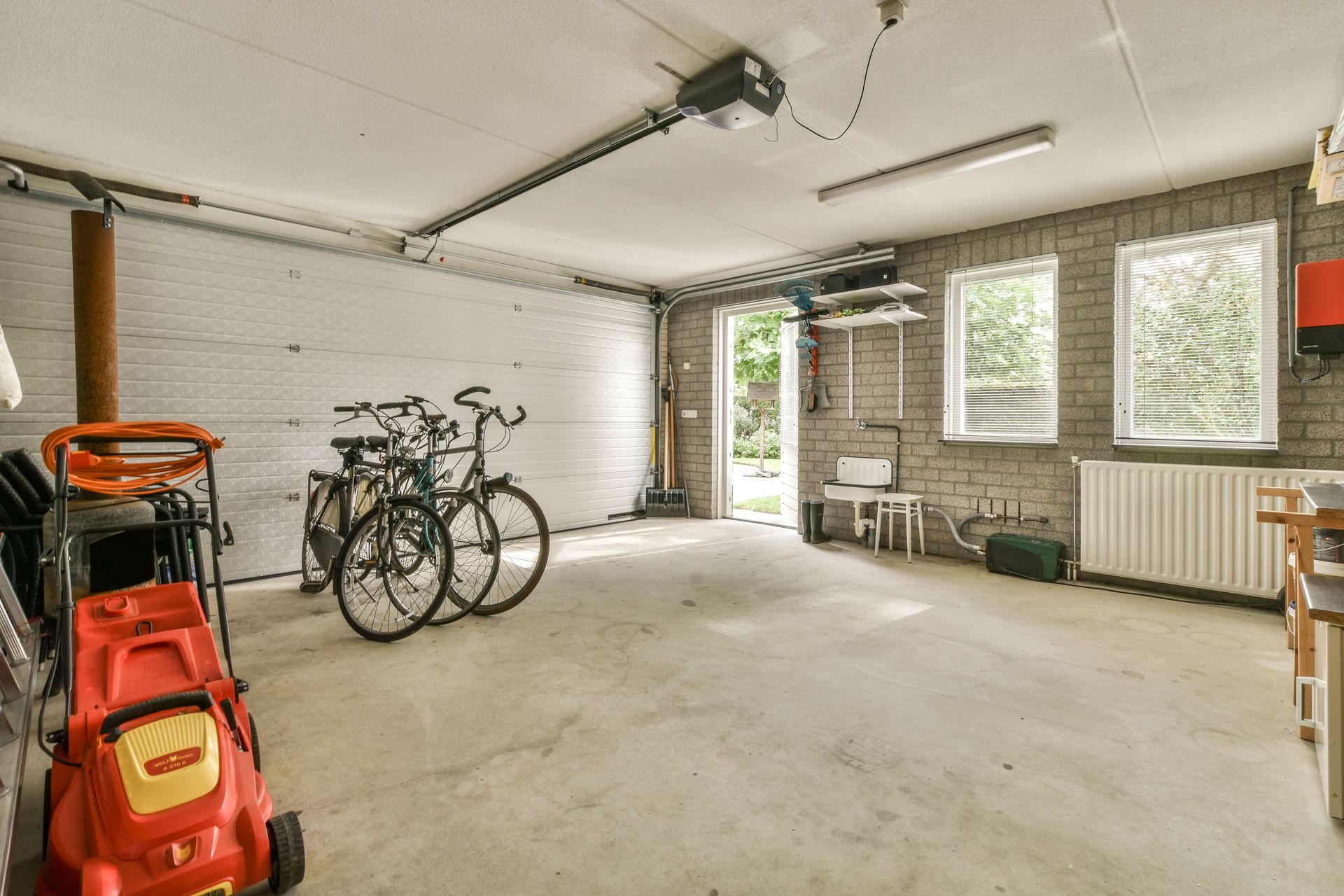 Garage interior with bicycles, lawnmower, and doorway to the outdoors. Brick and white painted walls, concrete floor.