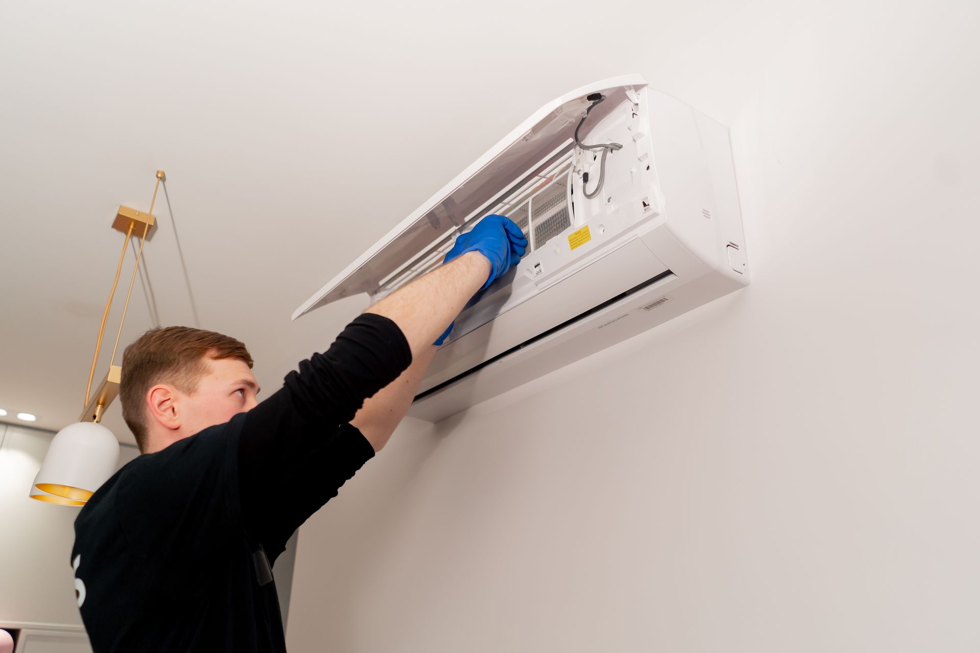 Person in blue gloves cleaning a wall-mounted air conditioner. They are holding open the top panel indoors.