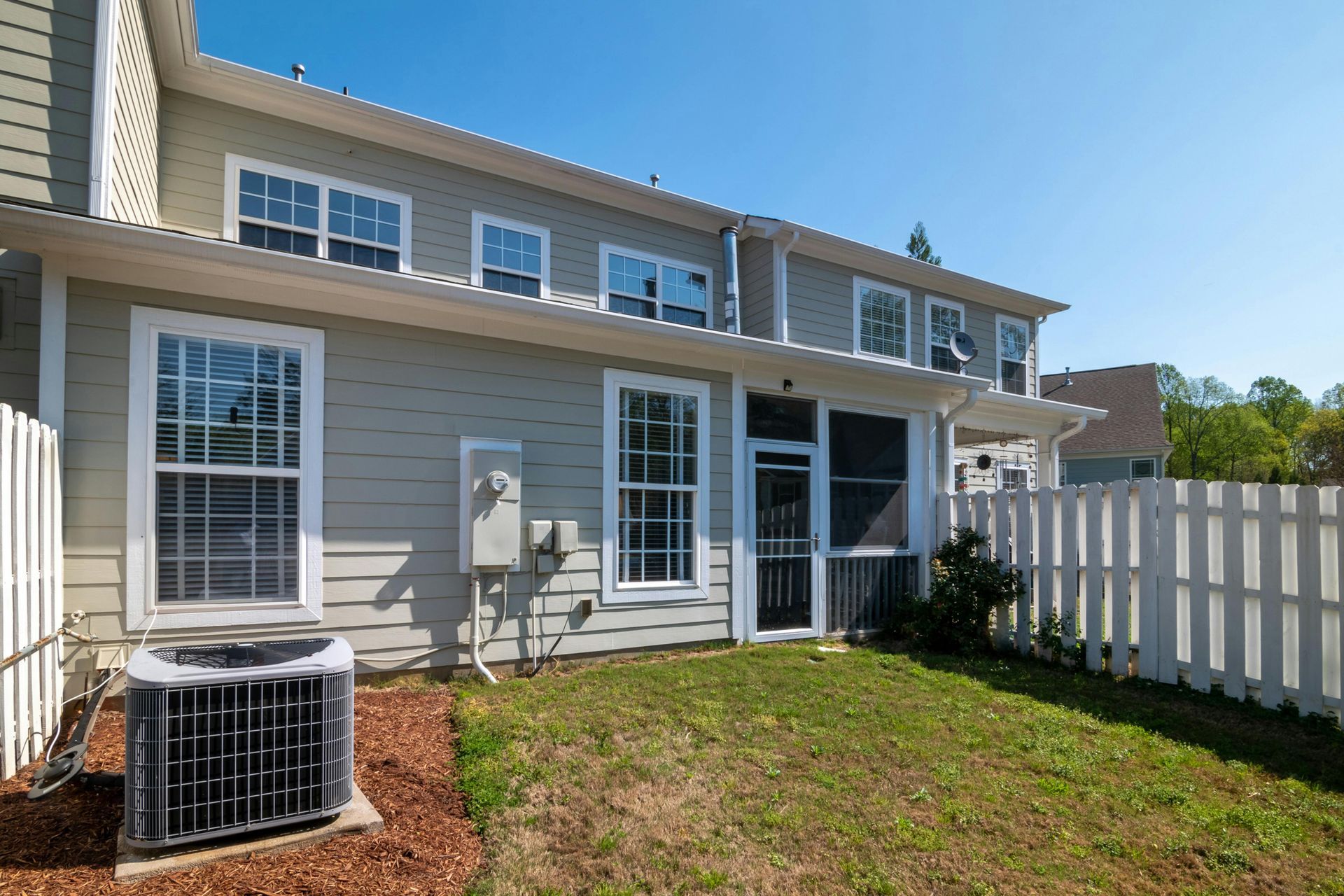 Backyard of a light gray townhome with a white fence, grass, and air conditioning unit.