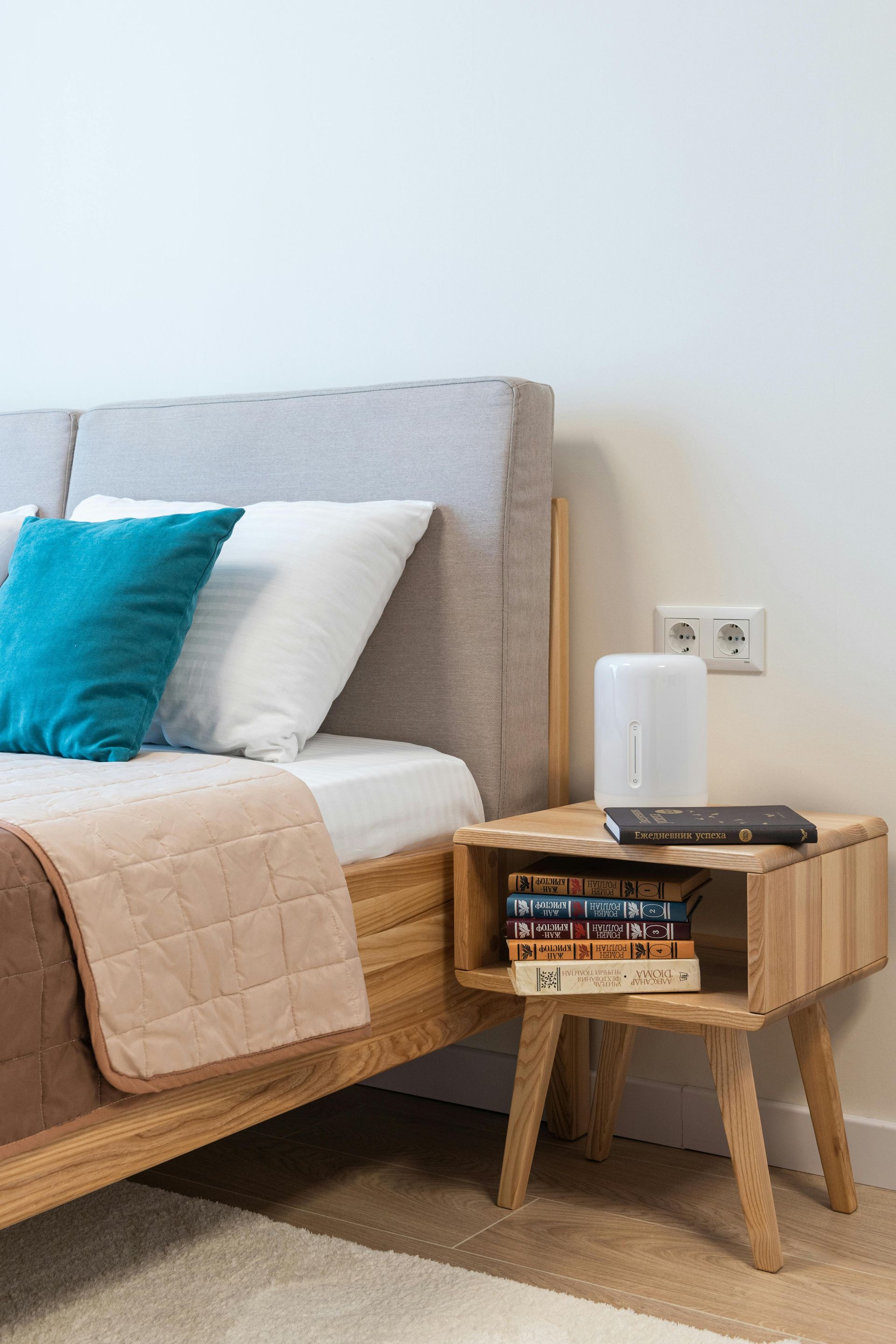 Bed with light wooden frame, pillows, blanket.  Nightstand with books, lamp, outlet.