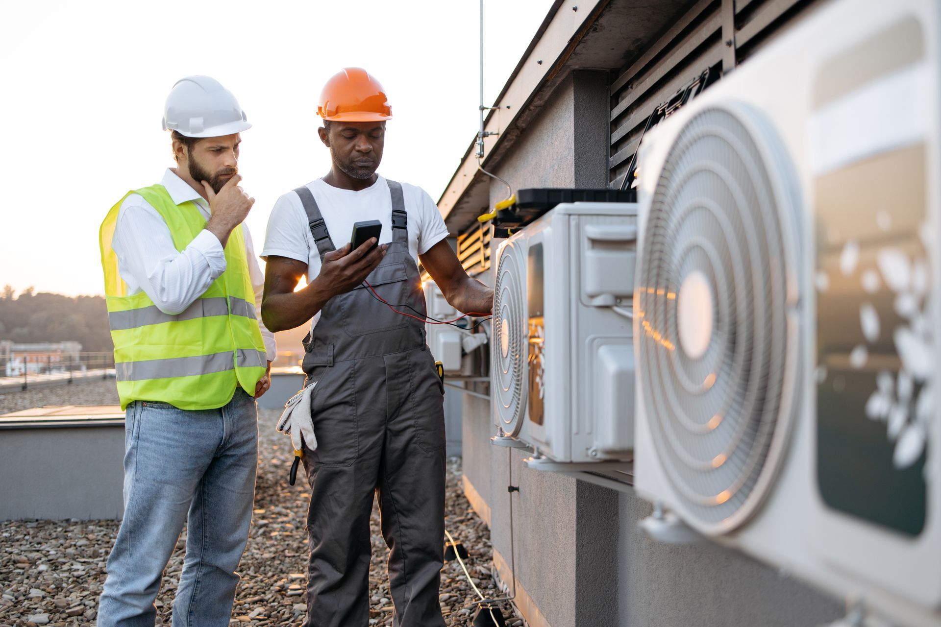 Two HVAC technicians on a rooftop, examining air conditioning units. One looks at a phone, the other observes.