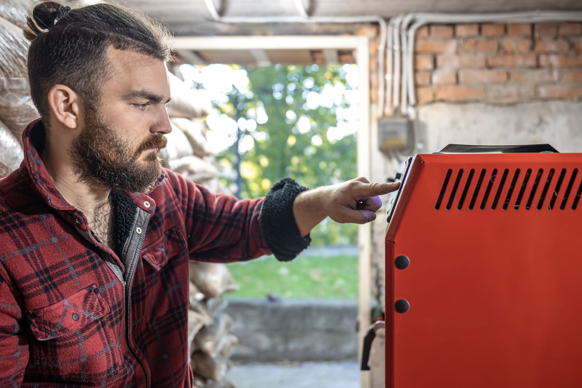 Man in red plaid shirt, presses a button on a red boiler. Interior setting.