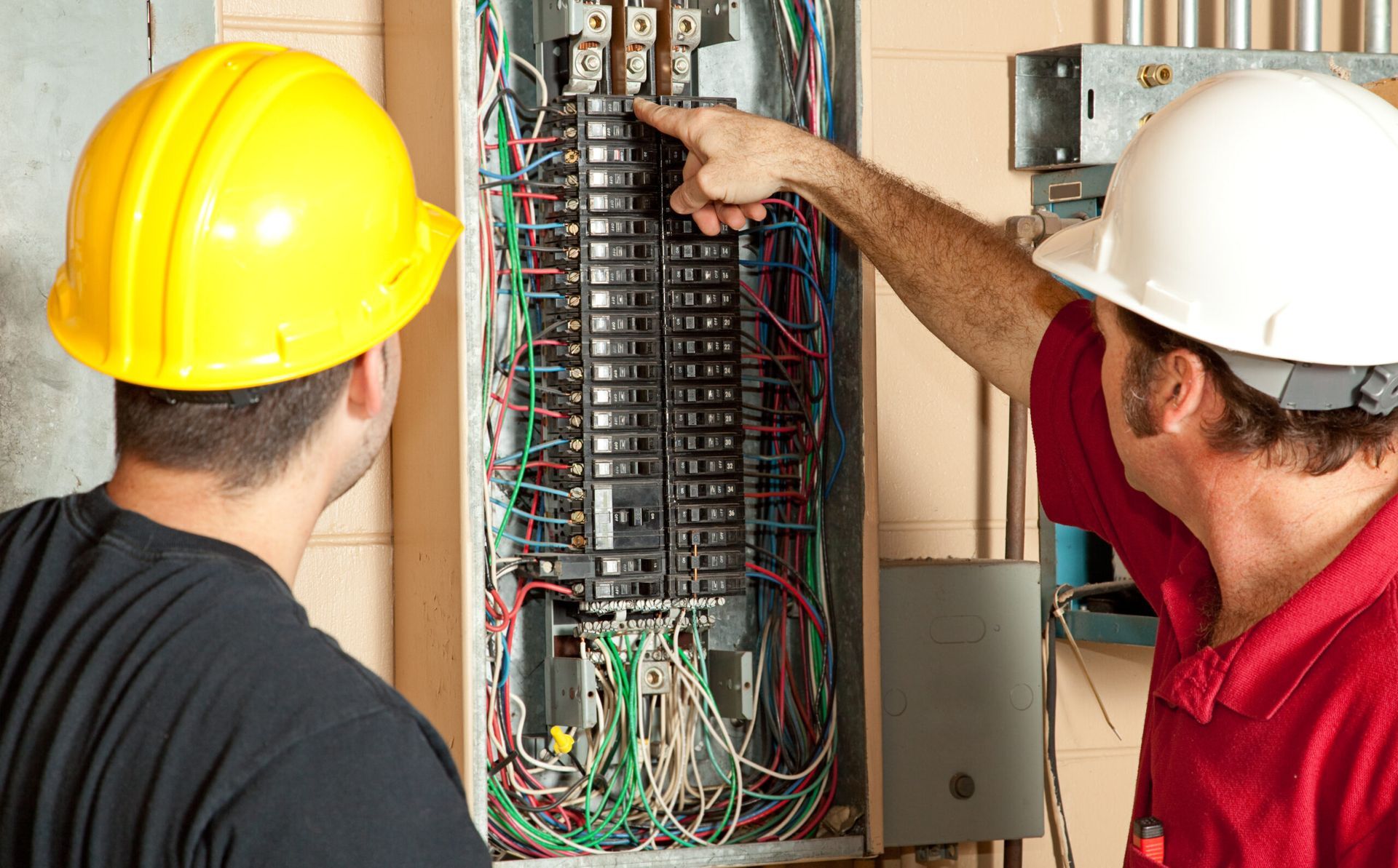 Two men wearing hard hats are working on an electrical panel.