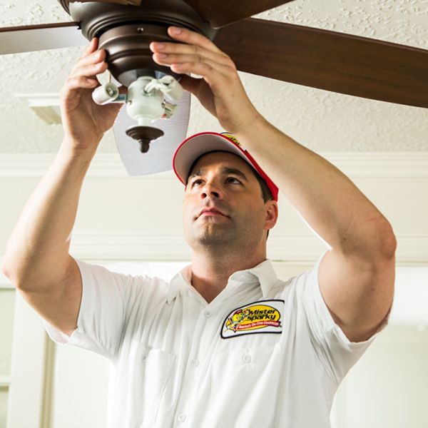 A man is fixing a ceiling fan while wearing a white shirt with a yellow sticker that says ' jackson '