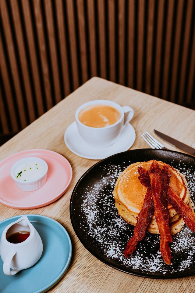 Panqueques con tocino y azúcar en polvo en un plato negro, café, almíbar y crema en una mesa.