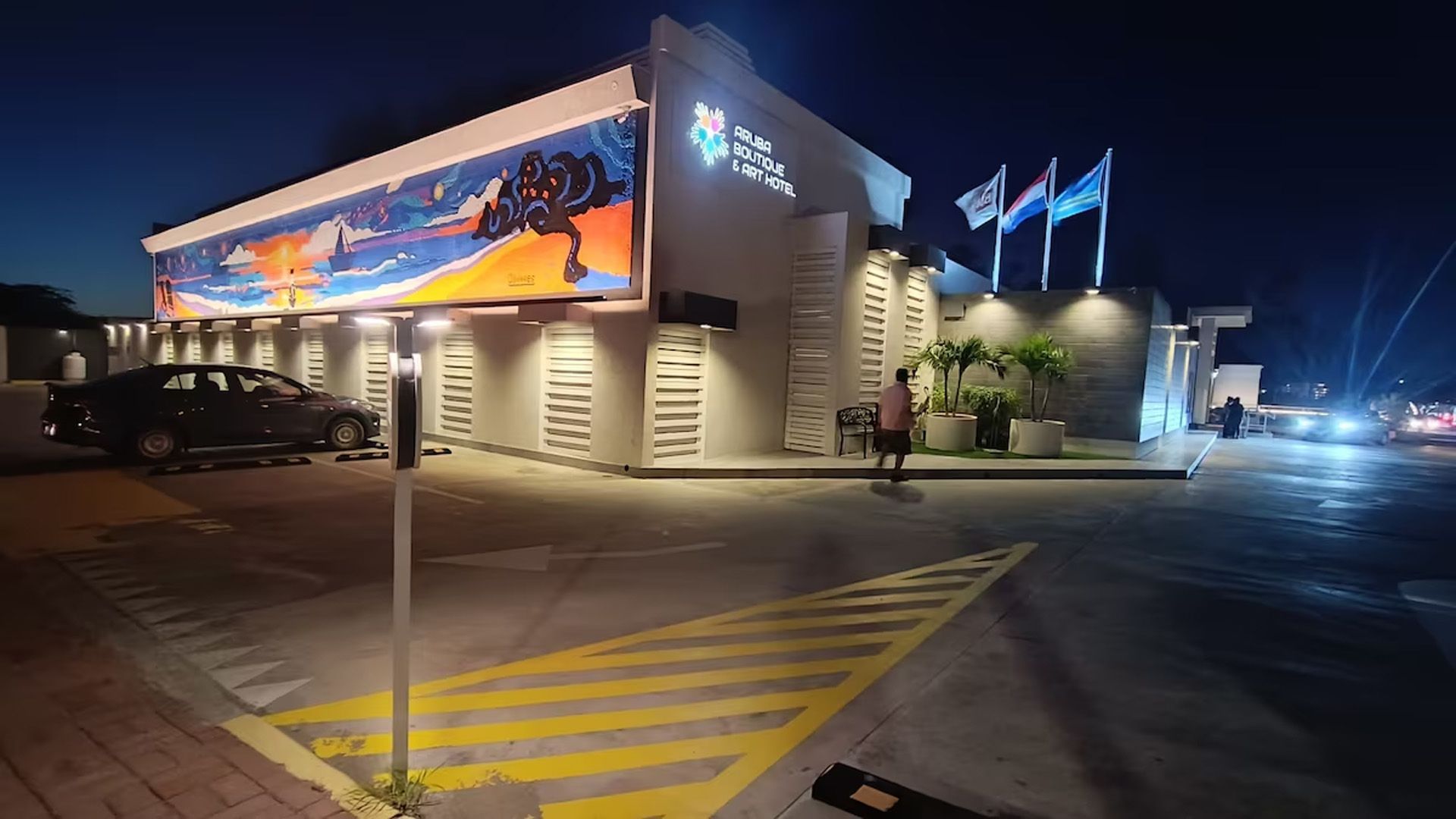 Exterior de un edificio brillantemente iluminado por la noche con un mosaico, banderas y un automóvil en el estacionamiento.