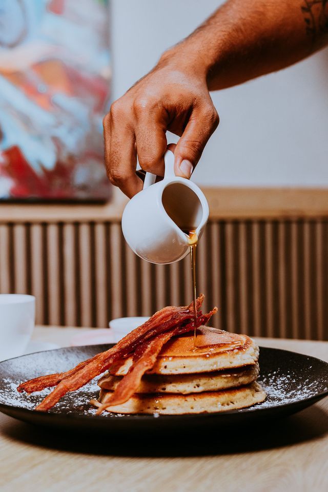 Persona vertiendo almíbar sobre panqueques cubiertos con tocino, servidos en un plato negro.