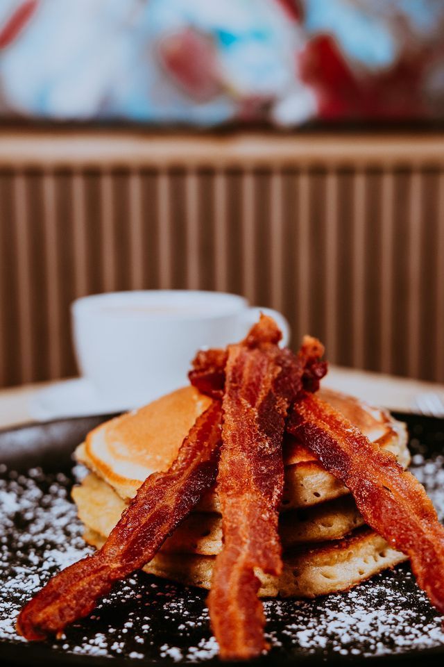 Panqueques apilados con tocino crujiente, espolvoreados con azúcar en polvo y una taza de café en el fondo.