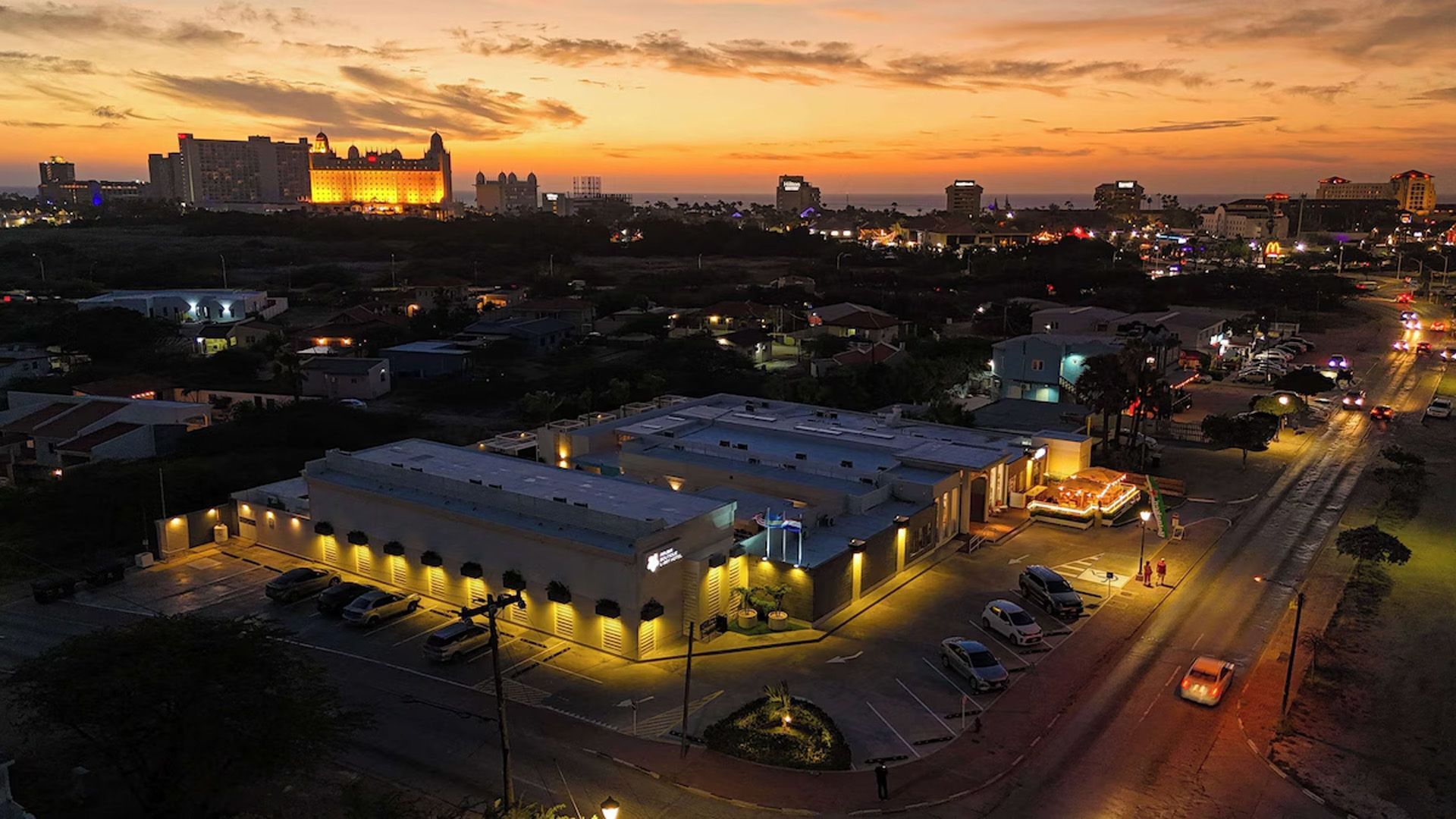 Vista aérea nocturna de un edificio iluminado con paisaje urbano circundante y una colorida puesta de sol.