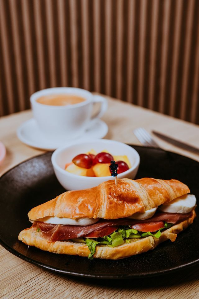 Sándwich de croissant con bol de fruta y café en un plato negro, entorno de cafetería.