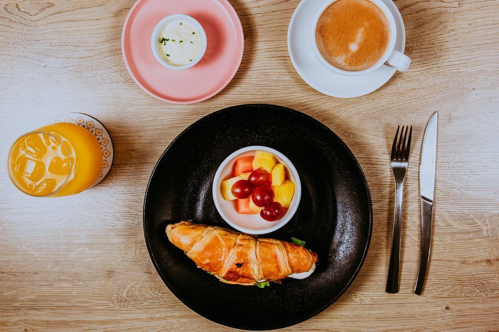 Plato de desayuno con croissant, fruta, café, mantequilla y naranja sobre mesa de madera.