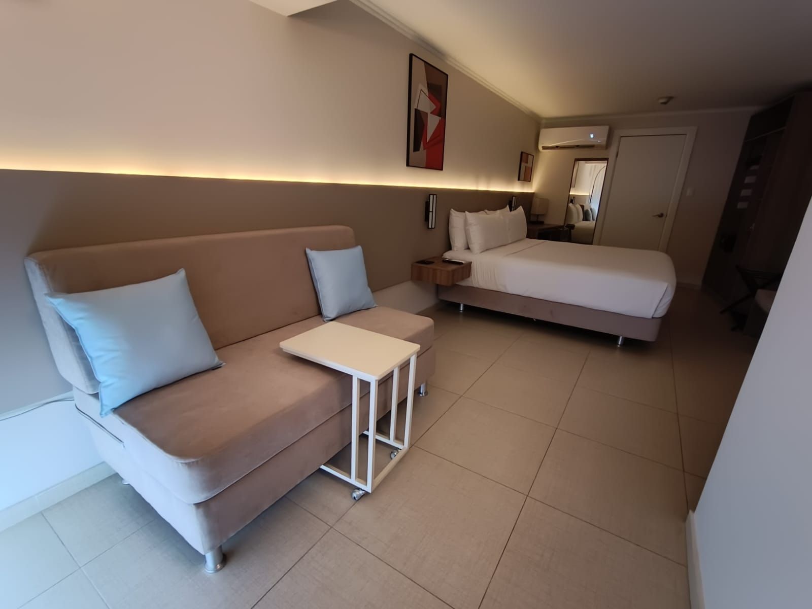 Habitación de hotel con sofá, cama y mesa pequeña; tonos neutros.