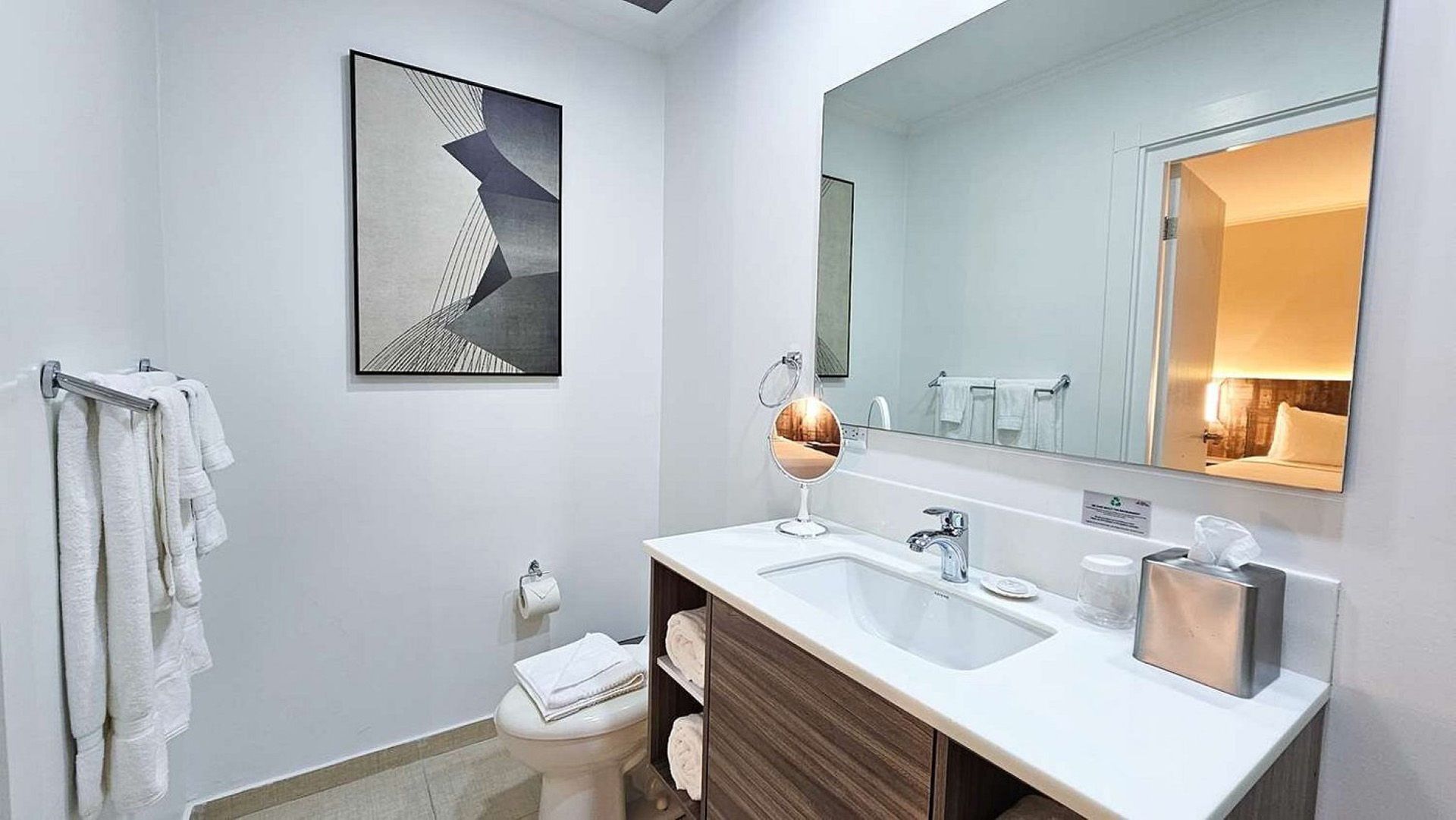 Baño moderno con encimera blanca, lavabo y espejo, toallas y vista a un dormitorio.