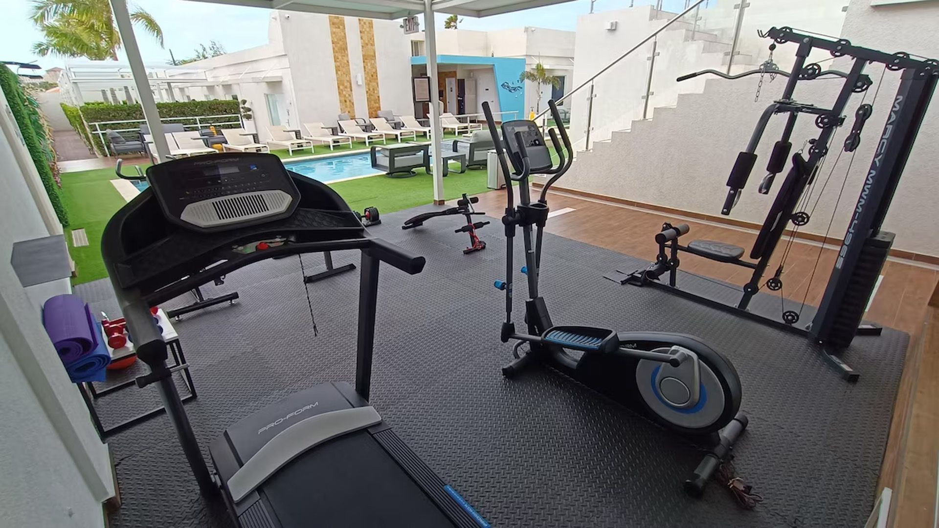 Gimnasio con cinta de correr, elíptica, máquina de pesas y piscina al fondo.