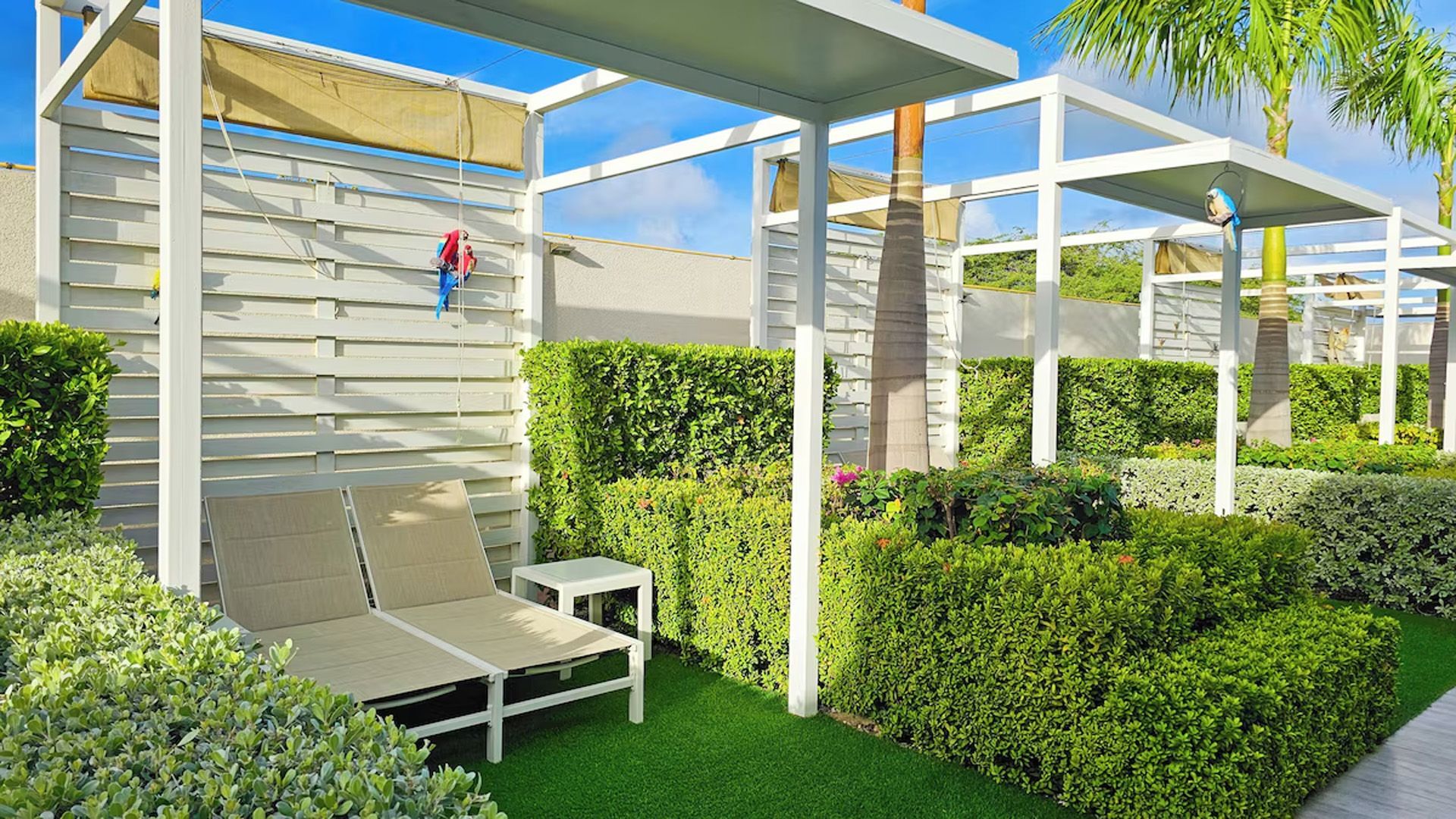 Tumbonas en cabañas privadas con setos y césped verde bajo pérgolas blancas en un entorno soleado al aire libre.