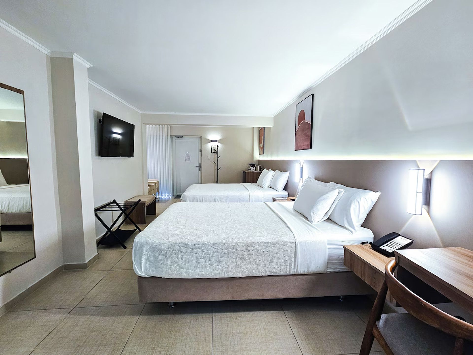 Habitación de hotel con dos camas, colores neutros, decoración moderna, escritorio de madera, silla y TV.