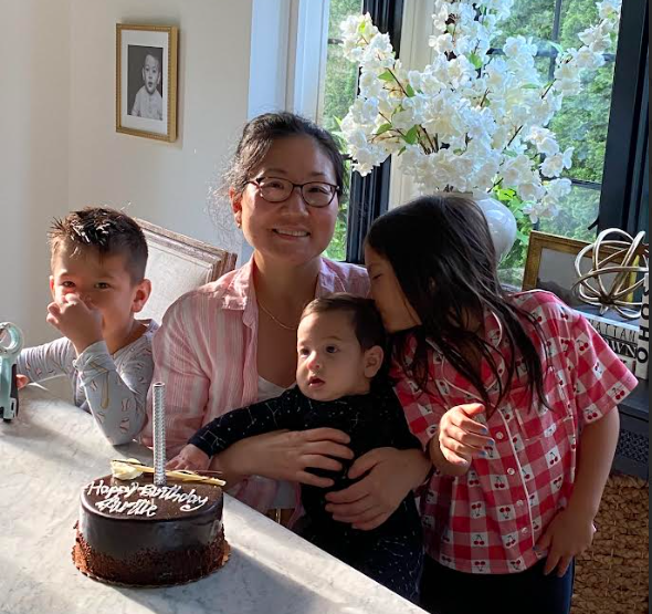 Woman with glasses smiles, holding a baby, surrounded by three children near a birthday cake. Bright, natural light fills the room.