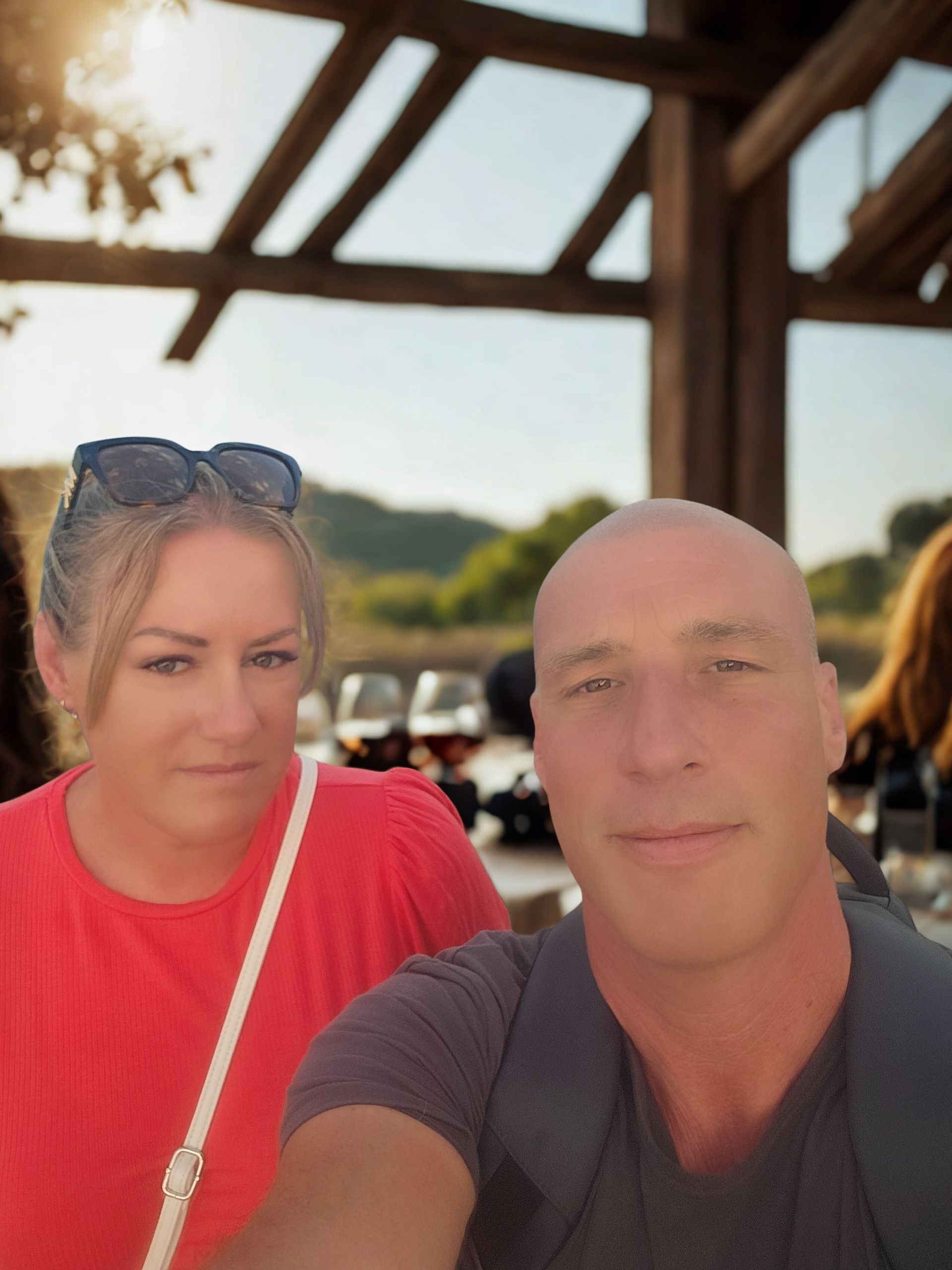 Vrouw en man aan een tafeltje buiten; vrouw in rode top, man in grijze. Zonlicht, wijnglazen zichtbaar.