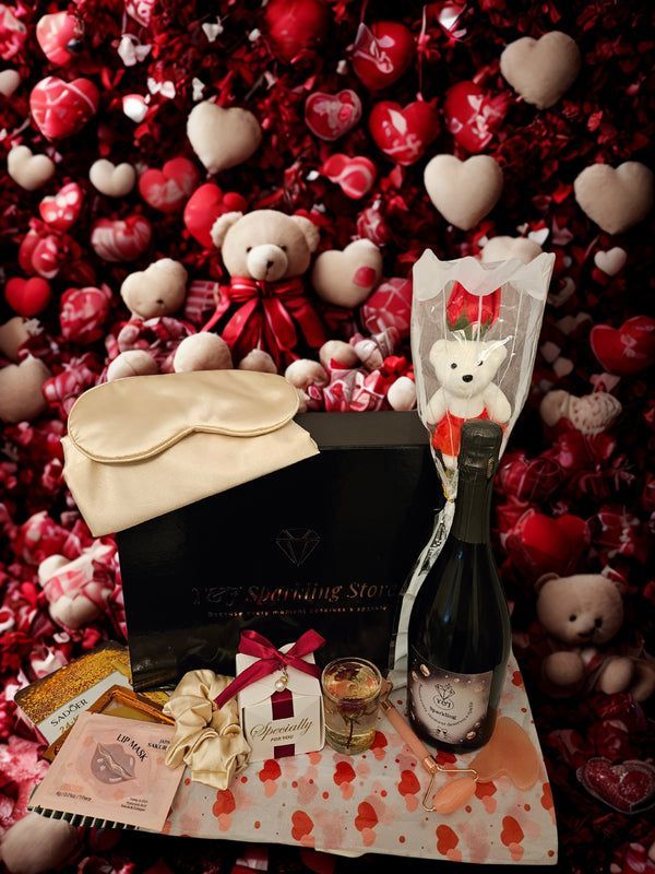 Valentijnsdag cadeauset: berenboeket, champagne, chocolaatjes, slaapmasker, kaars, tegen een rode hartvormige achtergrond.